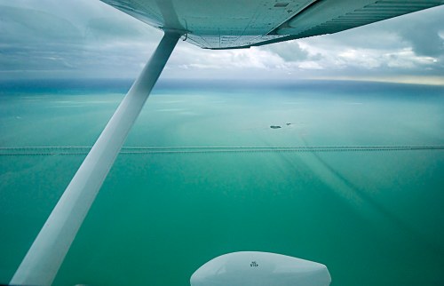 Aerial view from a plane showing a bridge over turquoise waters and a small island in the distance under cloudy skies.