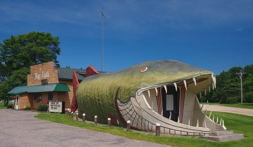 A large fish-shaped building serves as a quirky restaurant, set against a clear blue sky and green landscape.