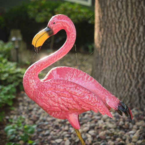 A pink flamingo garden decoration stands beside a tree, glistening in sunlight with a backdrop of greenery and stones.