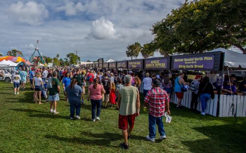 A bustling outdoor festival with food stalls and a large crowd enjoying the event under a partly cloudy sky.
