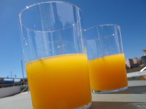 Two glasses of orange juice against a clear blue sky, with sunlight reflecting off the surface.