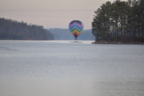 A colorful hot air balloon floats above a calm lake, surrounded by trees and a misty landscape.