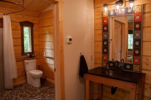 A rustic bathroom featuring wooden walls, a toilet, a sink with a mirror, and natural light from a window.