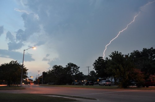 A street scene at dusk with a lightning bolt striking in the background, surrounded by dark clouds and trees.
