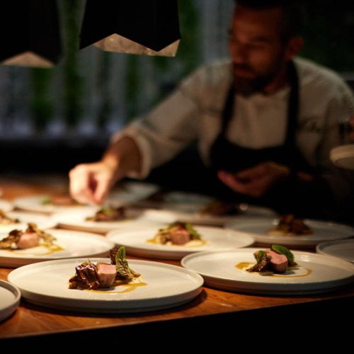 A chef carefully plating gourmet dishes in a dimly lit kitchen, with focus on the elegant presentation of the food.