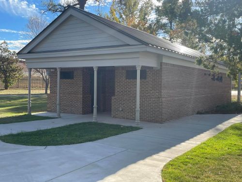 A brick building with a sloped roof, surrounded by grass and trees, featuring a concrete walkway.