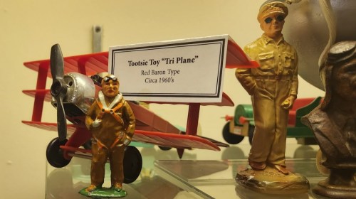 A red toy biplane labeled "Tootsie Toy 'Tri Plane'" next to a figurine of a pilot in a gold uniform, 1960s.