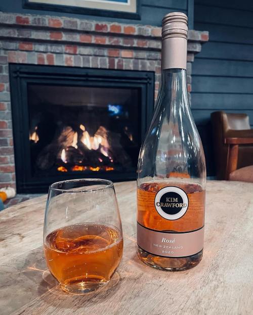 A bottle of Kim Crawford Rosé wine next to a glass filled with wine, with a cozy fireplace in the background.