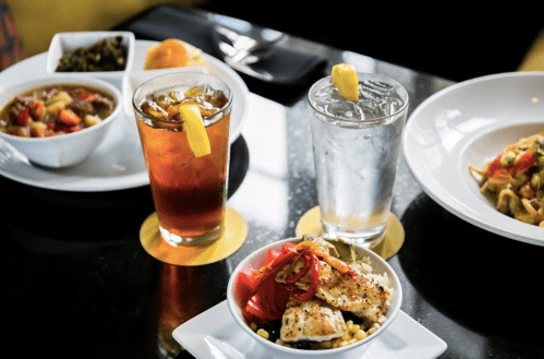 A table setting with two drinks, a bowl of grilled chicken with peppers, and two side dishes in white bowls.