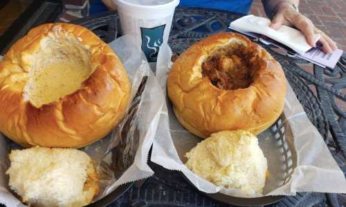 Two bread bowls filled with soup and stew, served with a side of bread, on a black metal table.