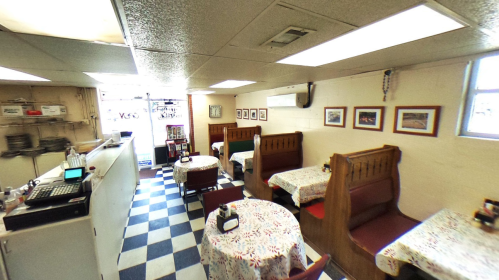 Cozy diner interior with checkered floor, wooden booths, and tables covered in floral tablecloths. Brightly lit and inviting.