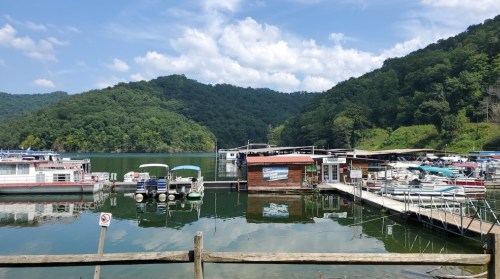 A serene lake scene with boats docked at a marina, surrounded by lush green hills and a blue sky with clouds.