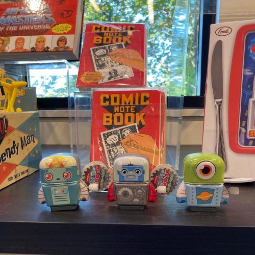 Three colorful robot figurines stand in front of comic note books, with a bright background of trees and display items.
