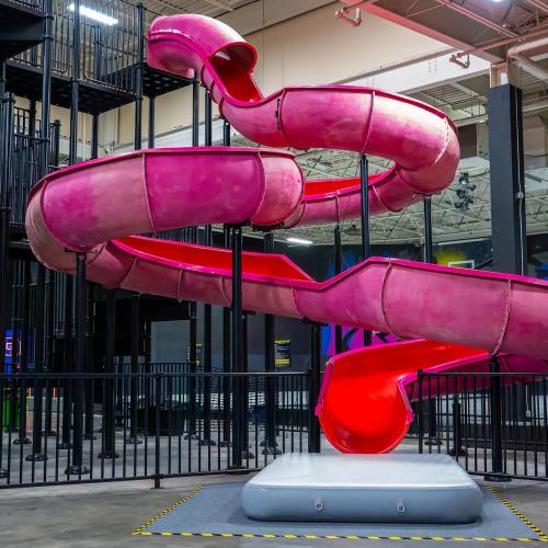 A bright pink water slide twists and turns in an indoor amusement area, surrounded by safety barriers.
