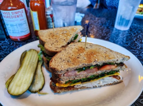 A sandwich with layers of lettuce, tomato, and cheese, served with pickles on the side, on a white plate.