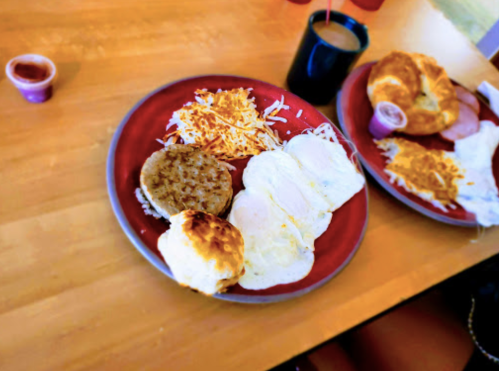 A red plate with two fried eggs, a biscuit, hash browns, a sausage patty, and a small cup of sauce.