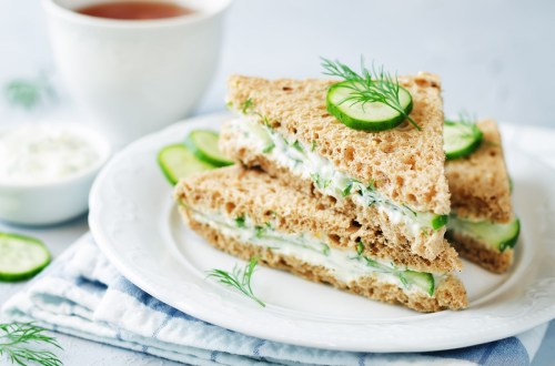 A plate with three cucumber sandwiches on whole grain bread, garnished with dill, alongside a cup of tea.