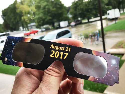 A hand holding eclipse glasses with a cosmic design, featuring the date "August 21, 2017" printed on them.