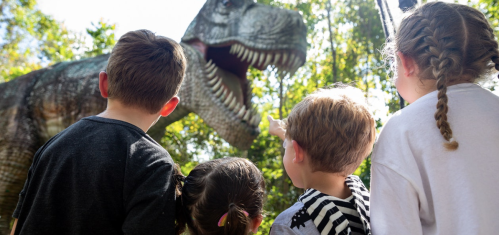 Four children gaze in awe at a large dinosaur statue in a forested area.