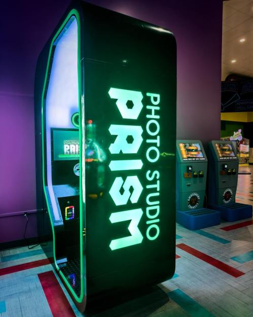 A brightly lit photo booth labeled "PRISM PHOTO STUDIO" in a colorful arcade setting.