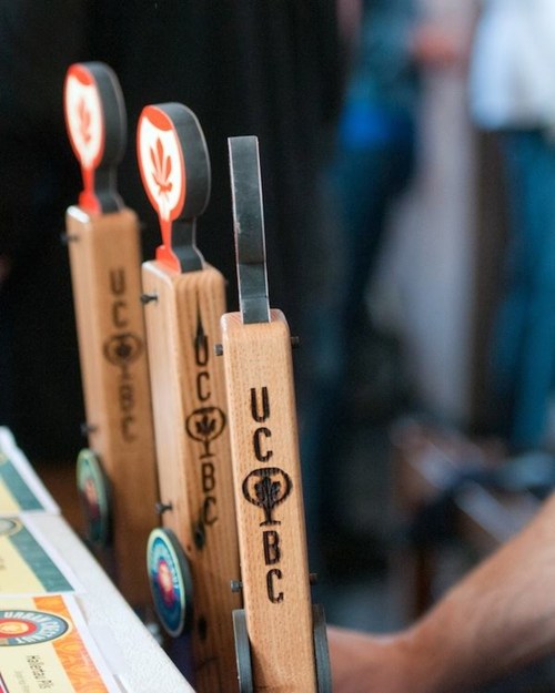 Three wooden beer tap handles with logos, featuring "UC" and "BC" engravings, set against a blurred background.