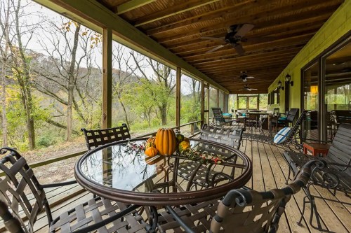 A cozy porch with a glass table, decorated with pumpkins and flowers, surrounded by trees and nature.