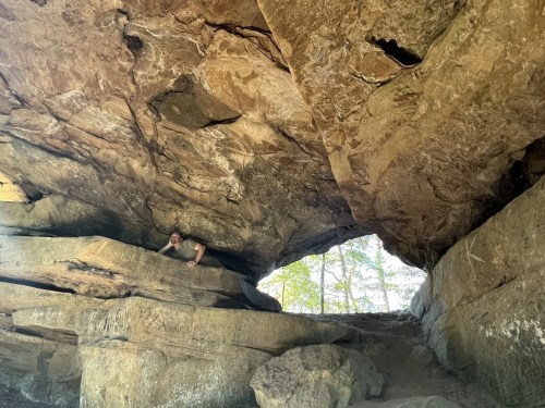 A person exploring a rocky cave with a large overhanging ceiling and natural light filtering in from an opening.