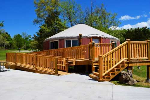 A wooden ramp leads to a round yurt surrounded by greenery under a clear blue sky.