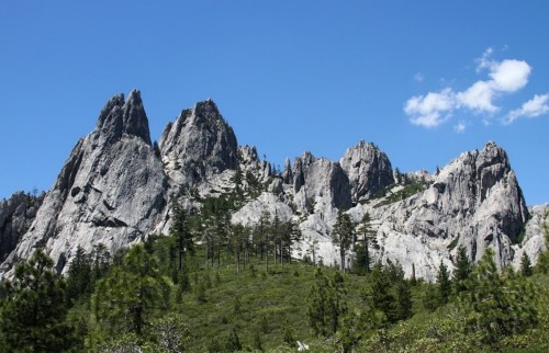 Majestic rocky peaks rise above a lush green landscape under a clear blue sky with a few scattered clouds.