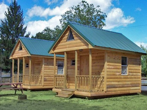 Two wooden cabins with green metal roofs, surrounded by grass and trees under a blue sky with clouds.