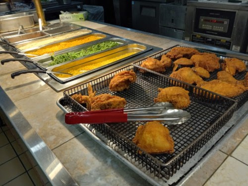 A buffet setup featuring fried chicken, green beans, and yellow rice in metal trays. Tongs are placed on the chicken.