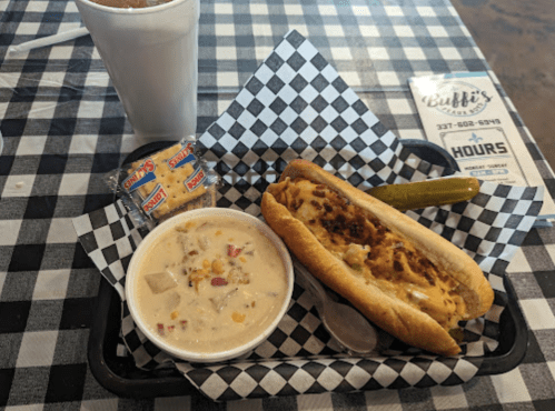 A hot dog topped with cheese and bacon, a bowl of chowder, a pickle, and condiments on a checkered tablecloth.
