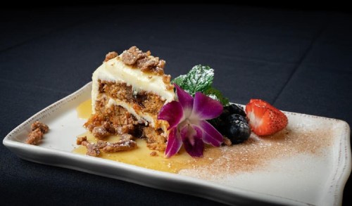 A slice of layered dessert topped with cream, nuts, and a flower, served with strawberries and blueberries on a plate.