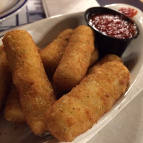 A dish of golden-brown mozzarella sticks served with a small cup of marinara sauce.