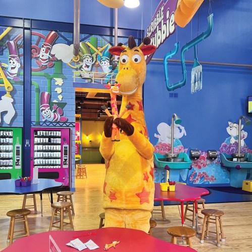 A cheerful giraffe mascot holds a toy in a colorful indoor play area with vending machines and playful decor.
