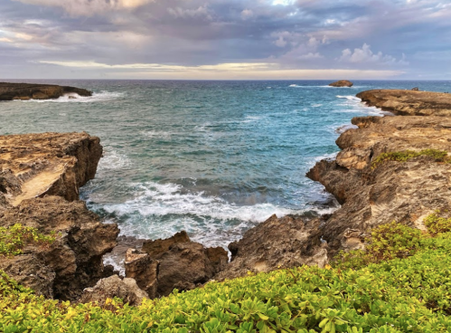 Coastal view with rocky cliffs, turquoise waves, and a cloudy sky in the background.