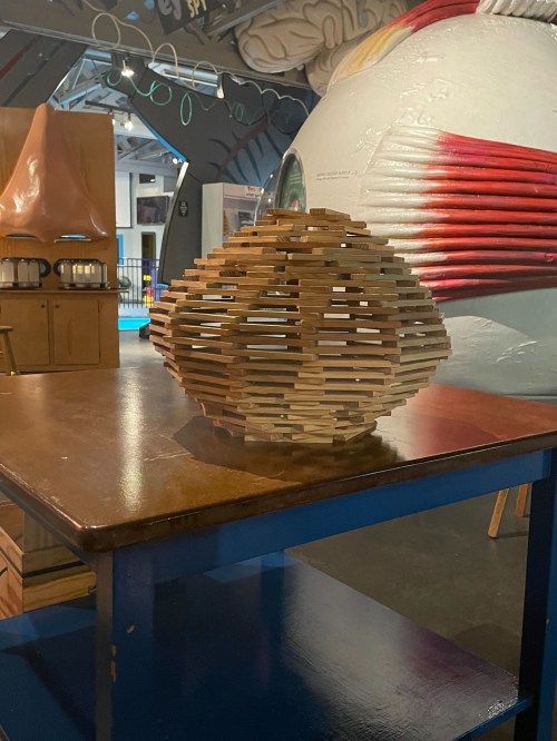 A wooden sculpture made of stacked pieces, resembling an orb, placed on a table in a colorful exhibit space.