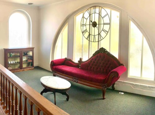 A cozy room featuring a red velvet sofa, a marble coffee table, and a large clock against arched windows.