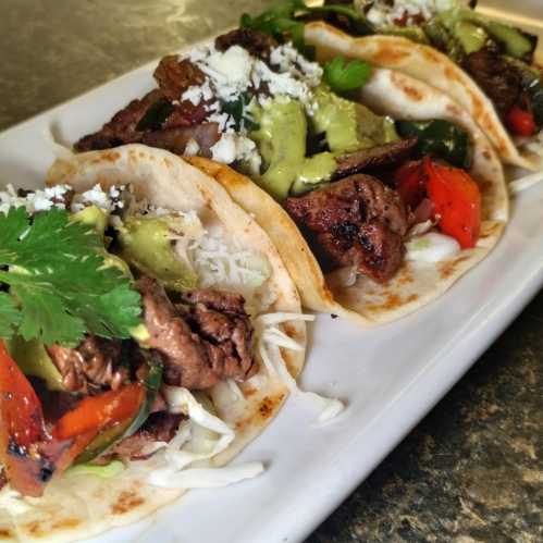 Three tacos filled with grilled beef, peppers, onions, avocado, cheese, and cilantro on a white plate.