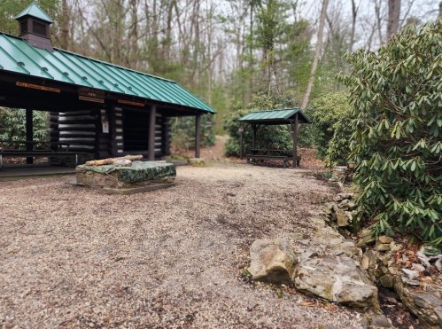 A rustic shelter with a green roof surrounded by trees and gravel, featuring a stone seating area and picnic tables.