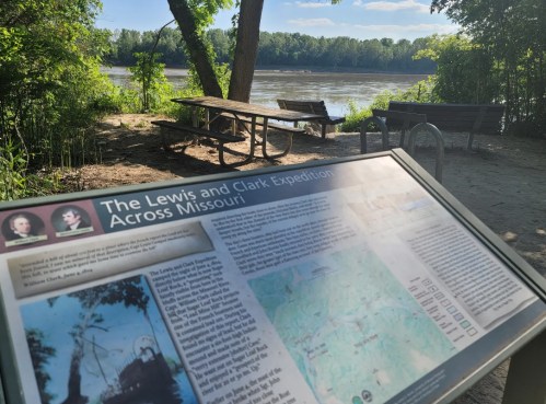 A sign about the Lewis and Clark Expedition overlooks a river, with benches and trees in the background.