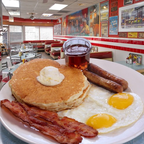 A plate of pancakes with butter, two eggs, sausage links, and bacon, alongside a glass of syrup in a diner setting.