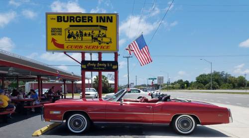 A red convertible parked outside Burger Inn, featuring a large sign and an American flag, on a sunny day.