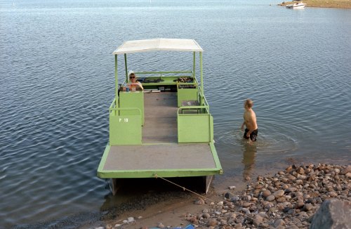 A green boat is anchored near the shore, with one person sitting inside and another standing in the water nearby.