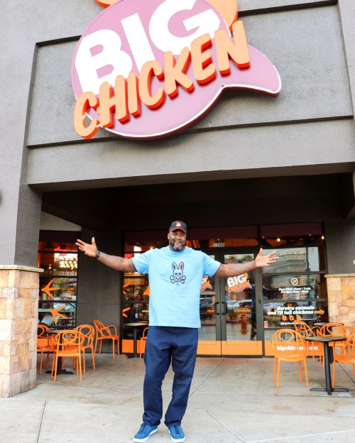 A man stands outside Big Chicken restaurant, smiling and welcoming guests with arms outstretched.