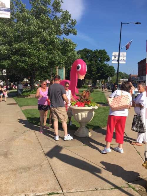 A group of people gather around a large pink flamingo decoration in a park on a sunny day.