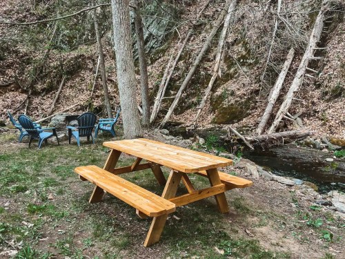 A wooden picnic table with benches near a stream, surrounded by trees and blue chairs in a natural setting.