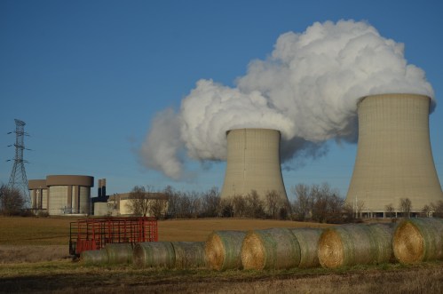Cooling towers release steam against a clear blue sky, with hay bales in the foreground and power lines in the background.