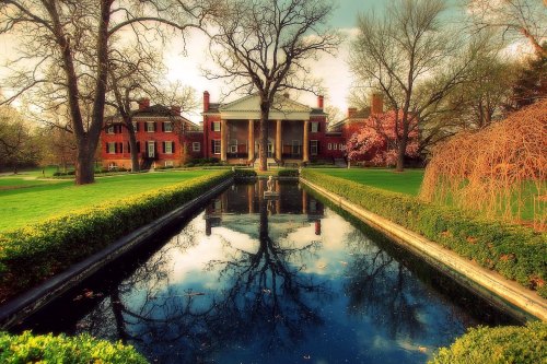 A grand mansion with columns reflected in a serene pond, surrounded by lush greenery and blooming trees.