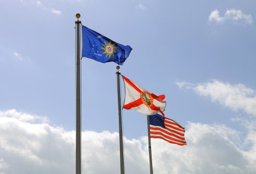 Three flags flying on poles: the Florida state flag, the U.S. flag, and a blue flag with a sun emblem against a cloudy sky.